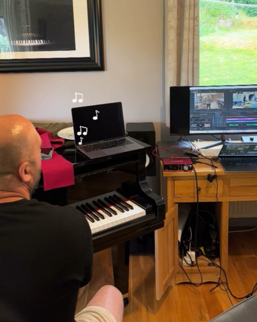 Image of man on piano facing computer with film edit playing while he composes music.